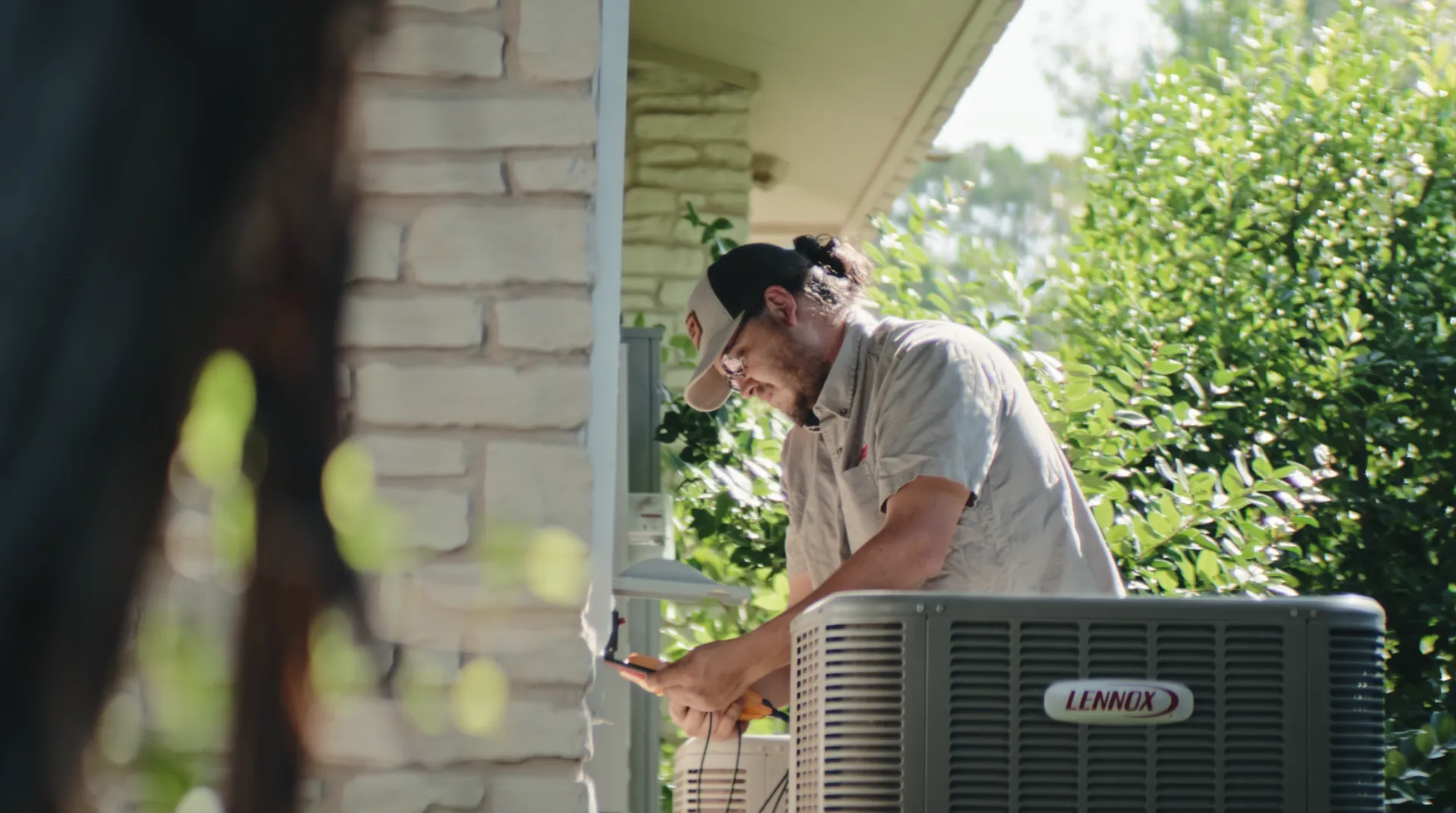 Technician in cap and glasses working on an outdoor Lennox air conditioning unit near a brick wall with green foliage in the background.