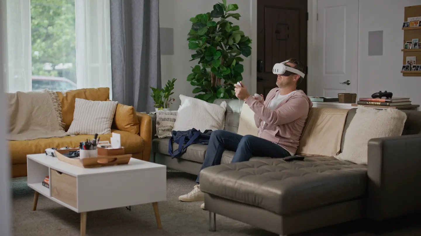 Man sitting on sofa playing with VR headset and controllers in a modern living room.