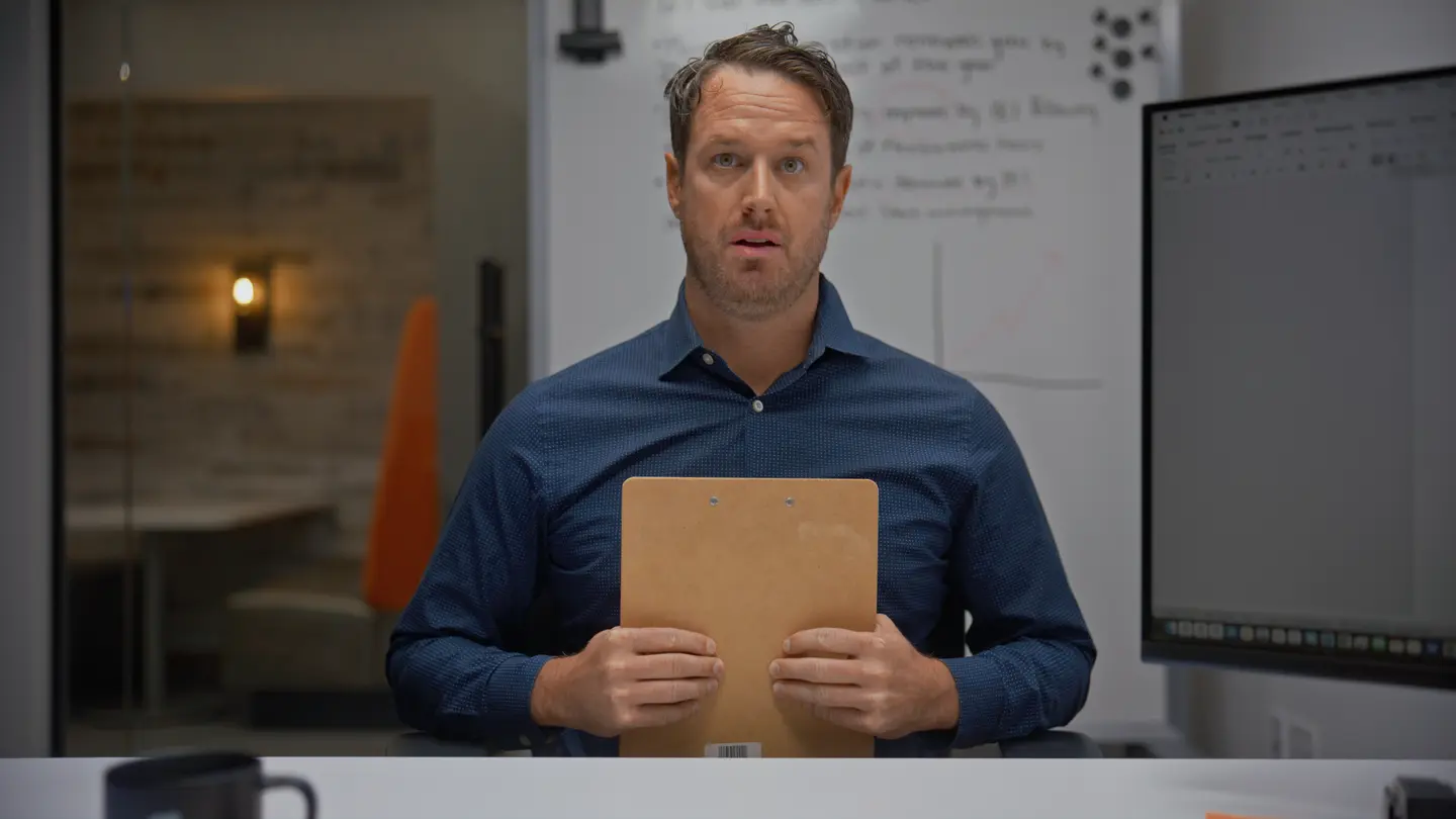 Man in blue shirt sitting at a desk holding a clipboard in an office with a whiteboard and screen behind.