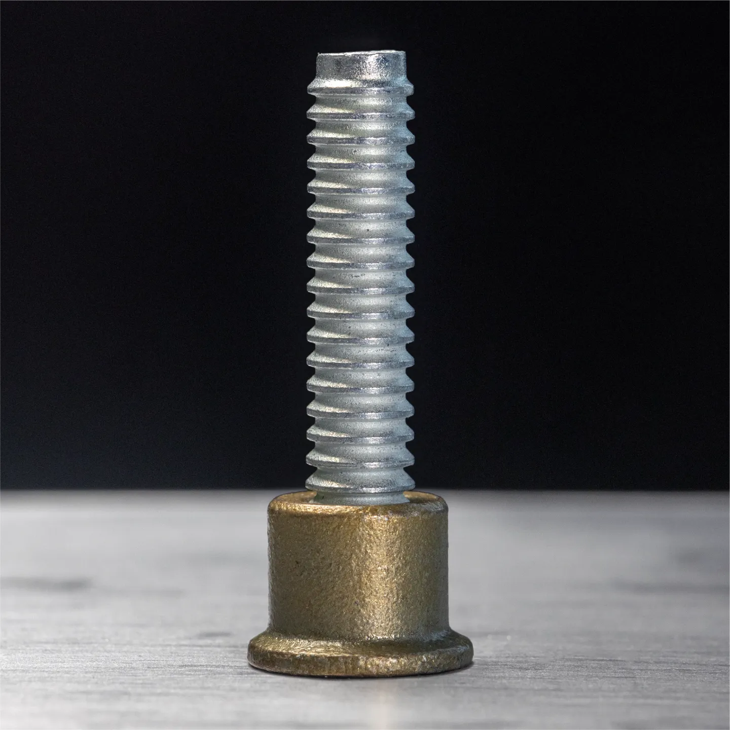Close-up of a silver threaded screw partially inserted into a bronze cylindrical base on a gray surface.