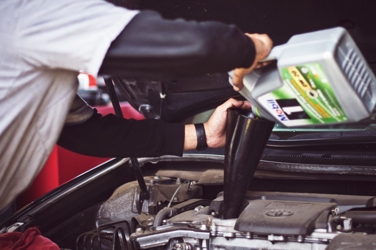 Technician pours motor oil into engine through funnel under open hood, performing routine oil change and maintenance service.