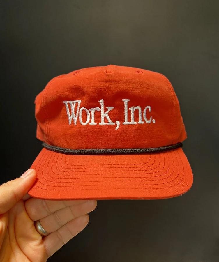 A hand holding an orange Work, Inc. cap.