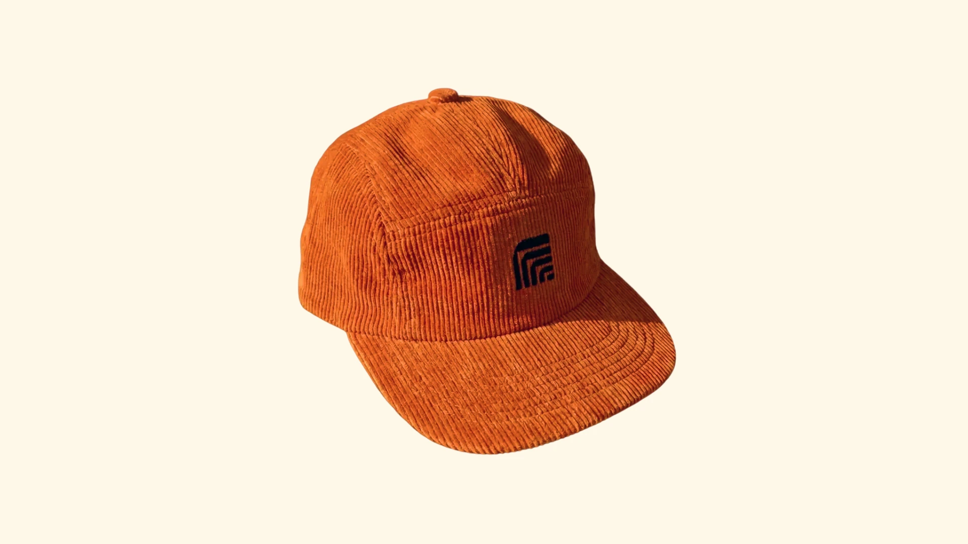 Orange corduroy cap embroidered with the black Rig symbol, shown on a warm cream background.