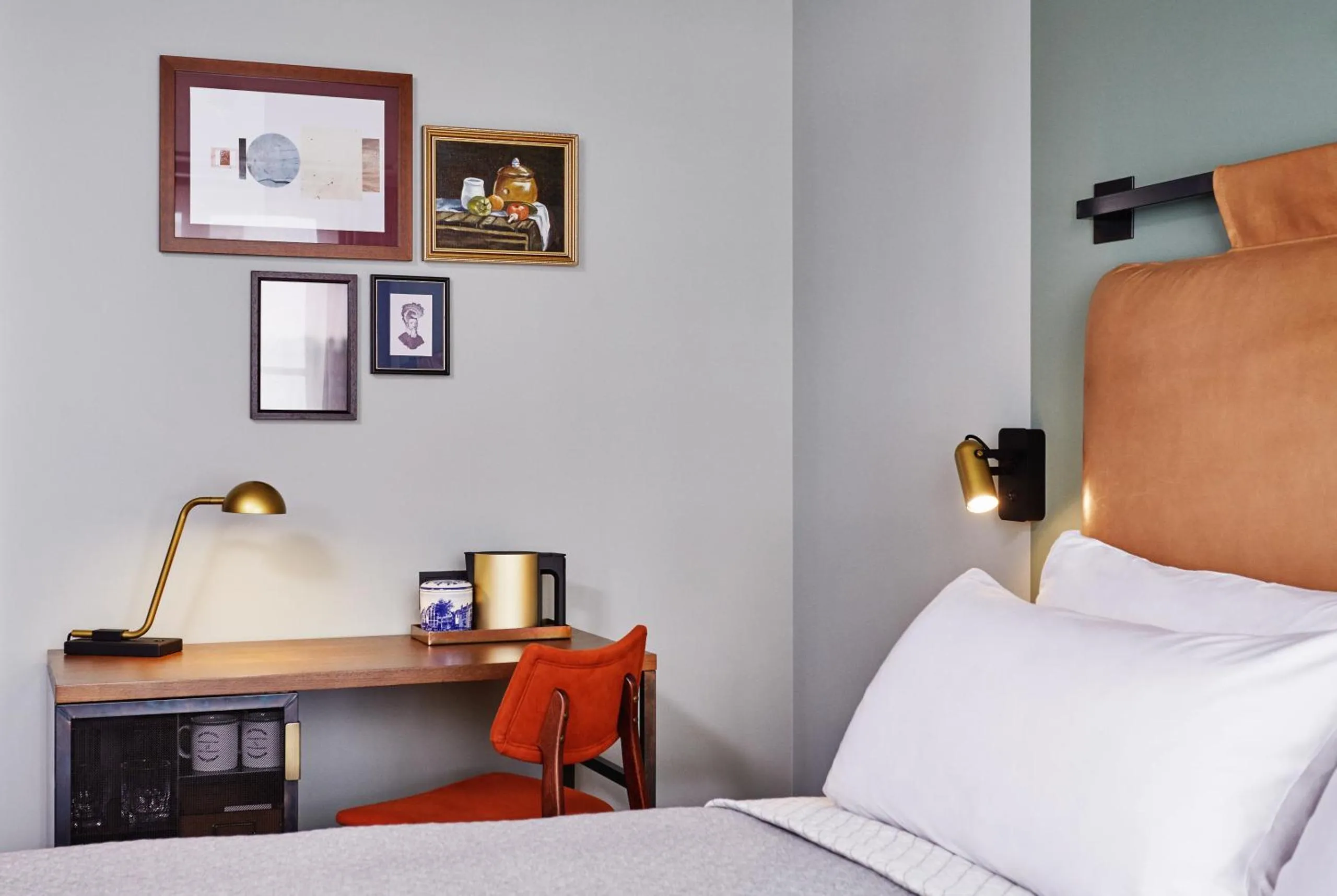 The Hoxton Amsterdam guest room with wooden writing desk, brass lamp, rust chair, gallery wall and tan leather headboard