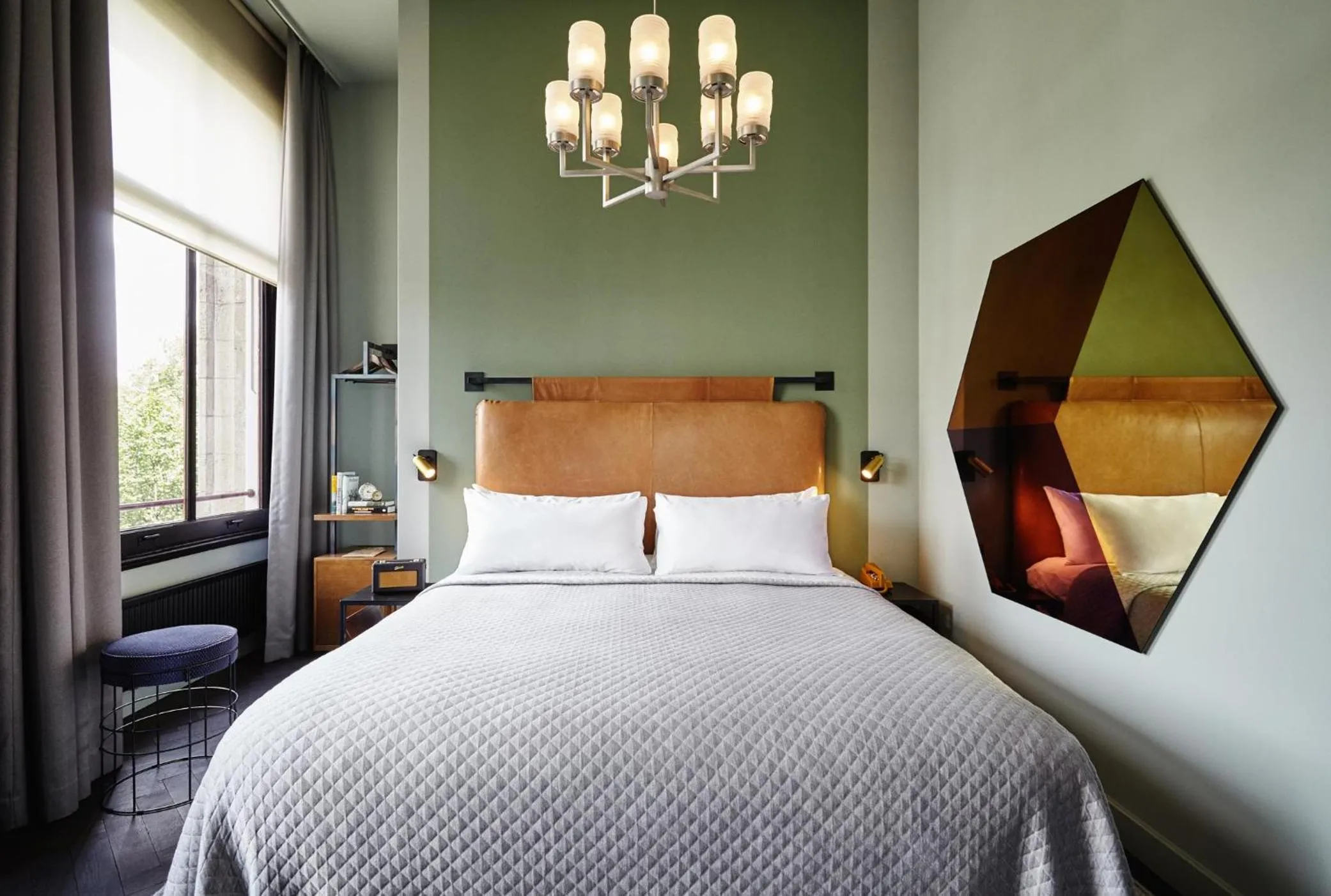 The Hoxton Amsterdam bedroom with sage green wall, tan leather headboard, hexagonal mirror and glass chandelier