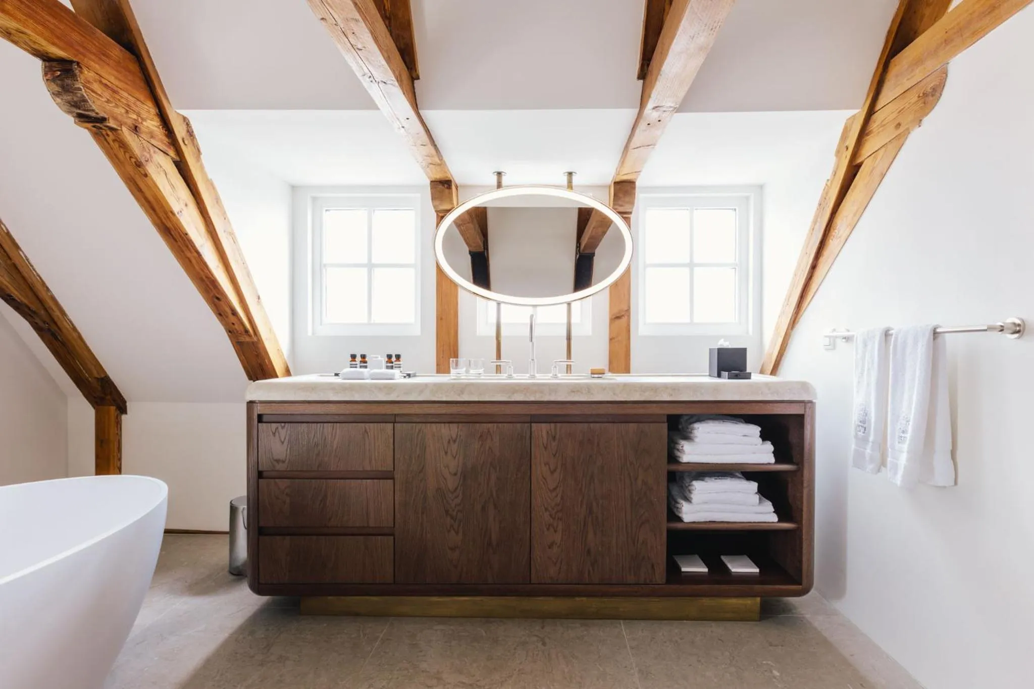 The Dylan Amsterdam bathroom with exposed timber beams, freestanding tub, walnut vanity and oval mirror