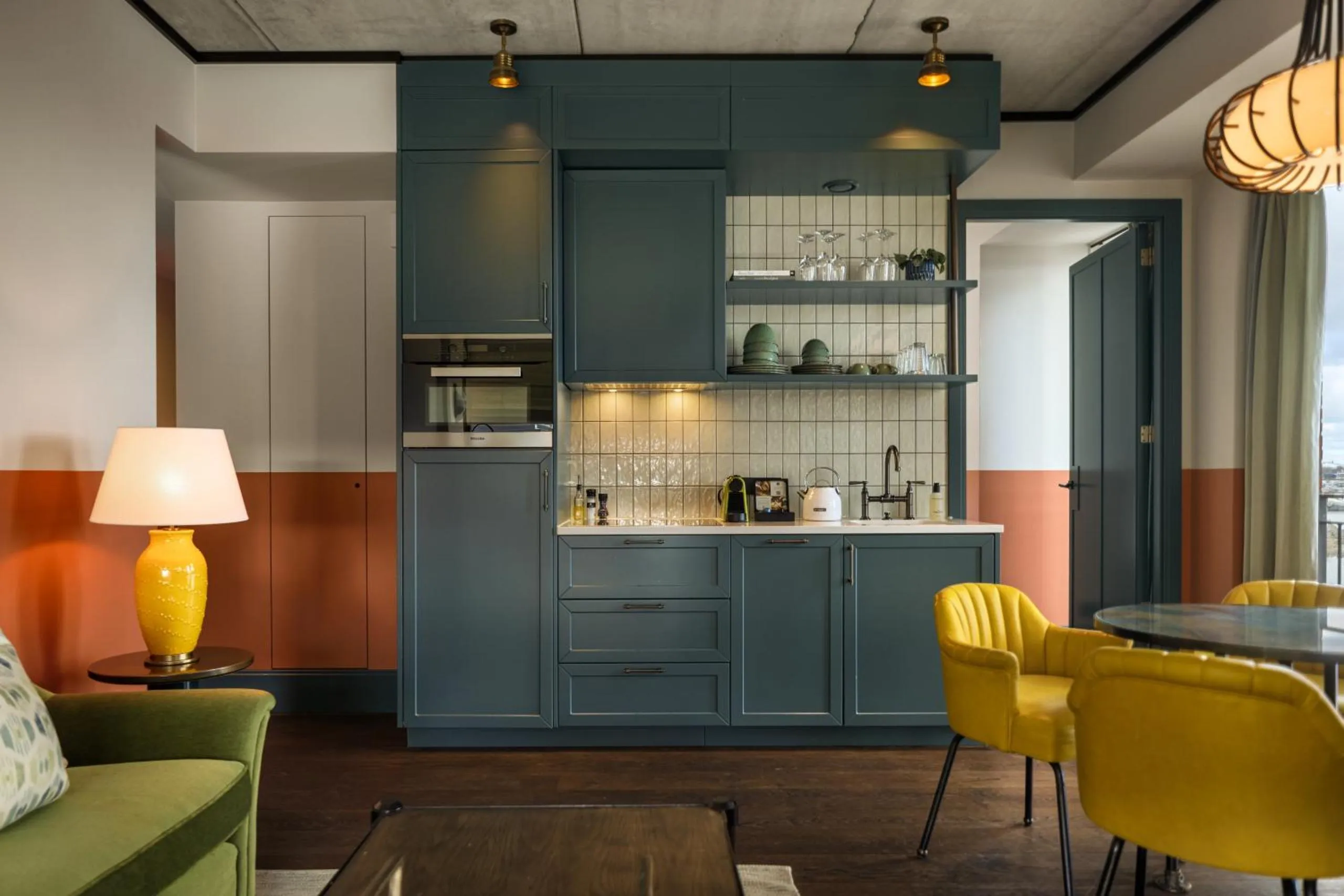 The July Boat & Co apartment kitchen with teal cabinetry, tiled splashback and mustard velvet chairs