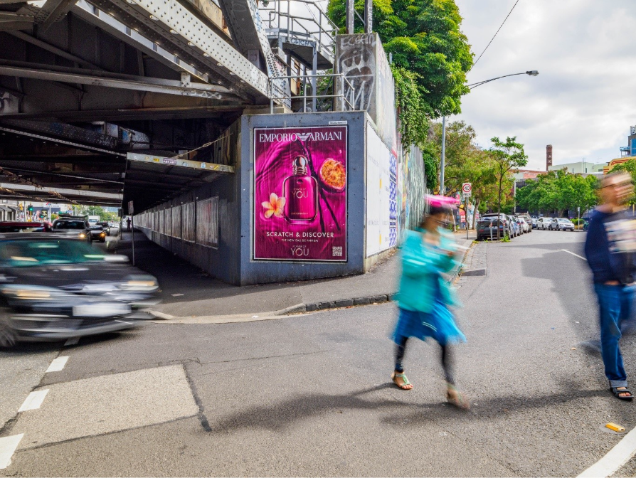 The image shows a street scene with a large, colorful advertisement for Revolution360 EMPORIO ARMANI on a bridge, with people walking and cars driving by.AI-generated content may be incorrect.
