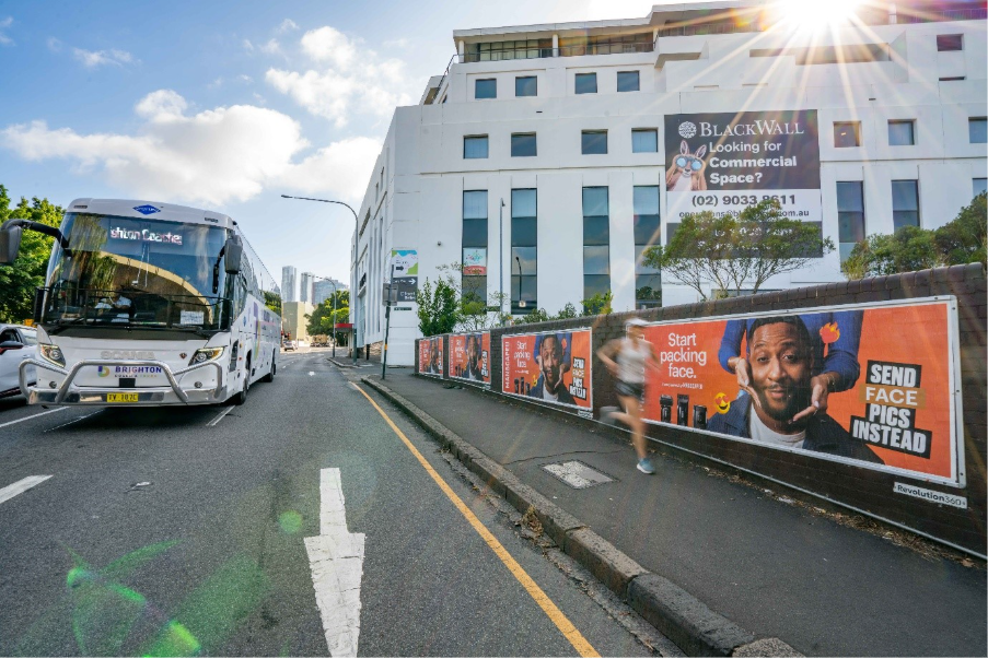 The image shows a bus parked on a street with a large advertisement on the side of the building, under a clear sky.AI-generated content may be incorrect.