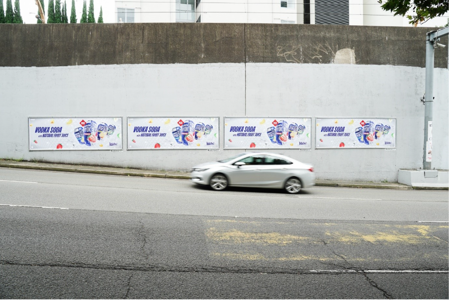The image shows a car parked on a street with a wall behind it displaying a series of colorful, stylized advertisements for a beverage, including Vodka Soda and natural fruit juices.AI-generated content may be incorrect.