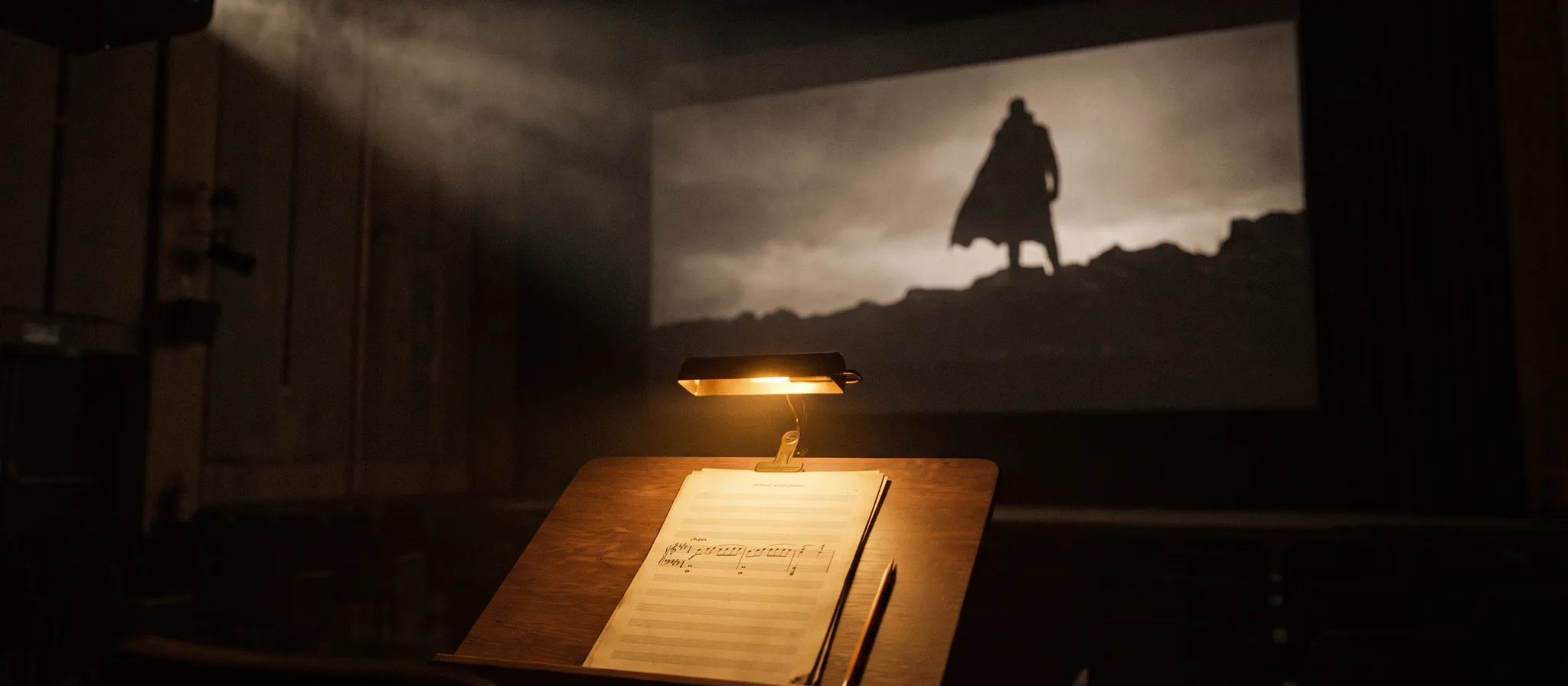 A lit music stand with a handwritten score in a dark scoring stage, a cloaked figure in silhouette on the cinema screen behind it