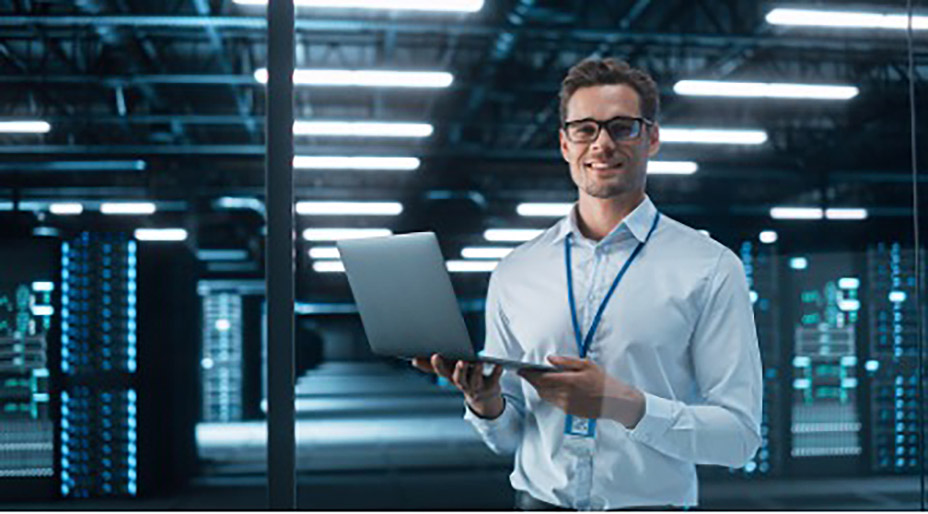 IT professional holding a laptop and smiling inside a modern data center with illuminated server racks in the background.