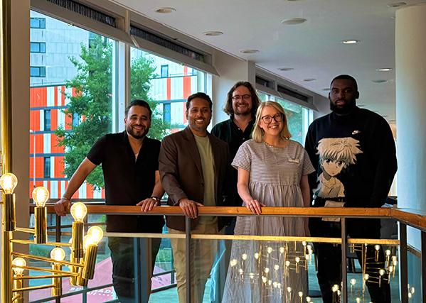 Five people standing side by side on an indoor balcony, smiling at the camera. Behind them are large windows showing modern buildings and greenery. The group stands in a well-lit area with decorative hanging lights in the foreground