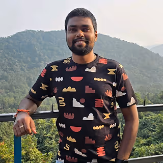 Smiling man with a beard wearing a patterned black T-shirt standing outdoors with green forested hills in the background.