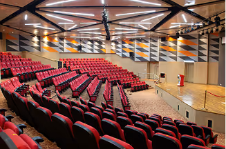 Indoor auditorium with red cushioned seats arranged in tiered rows and a wooden stage with a podium.