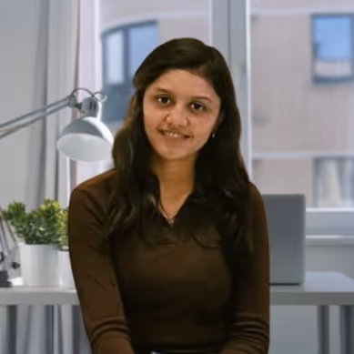Smiling woman with long dark hair wearing a brown top sitting indoors in front of a window and desk with a laptop and lamp.