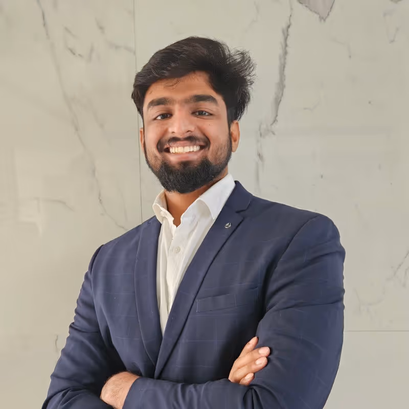 Smiling man with beard wearing a navy blue suit and white shirt standing with arms crossed against a marble wall.