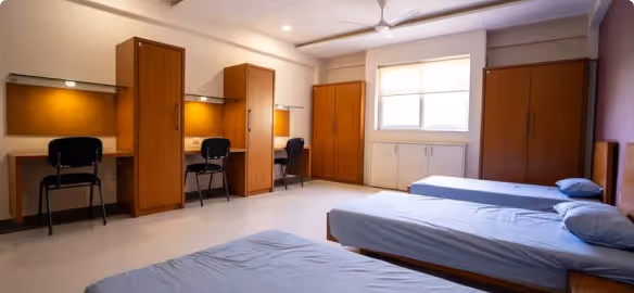Dormitory room with three single beds, study desks with chairs, and wooden wardrobes under warm lighting.