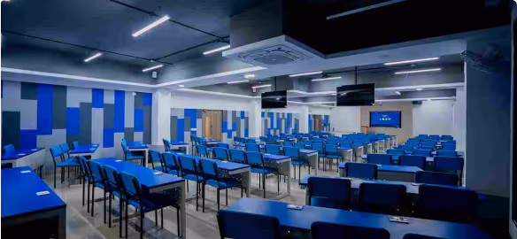 Modern classroom with rows of blue chairs and desks, colorful blue and gray wall panels, and ceiling-mounted air conditioning units.