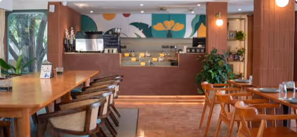 Modern cozy café interior with wooden tables, chairs, plants, and colorful wall art behind the counter.