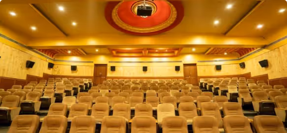 Empty auditorium with plush beige seats arranged in rows facing a stage, illuminated by warm ceiling lights.