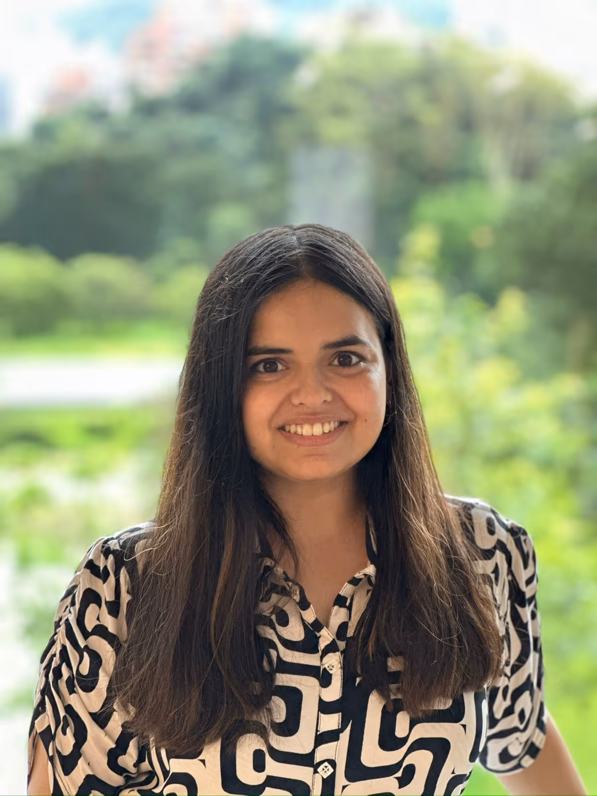 Smiling woman with long dark hair wearing a black and white patterned shirt standing outdoors with a blurred green background.