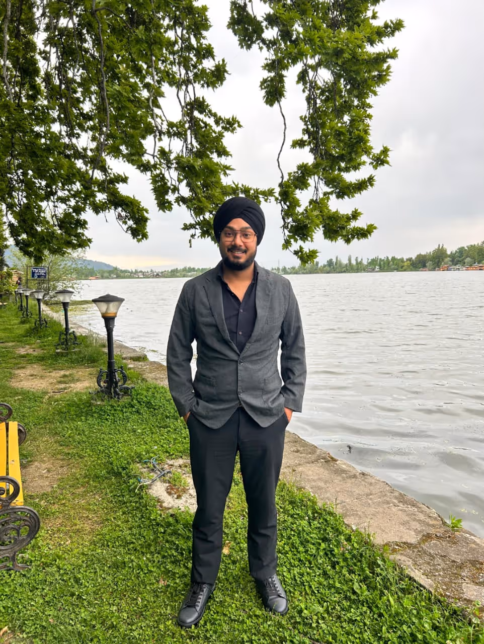 Man wearing a black turban, glasses, and dark suit standing on grass near a lake under green tree branches.