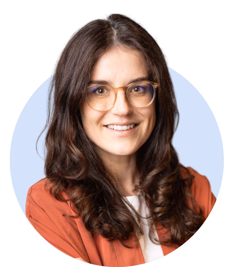 Smiling woman with long wavy dark brown hair wearing round glasses, a rust-colored jacket, and a white top against a light blue circular background.