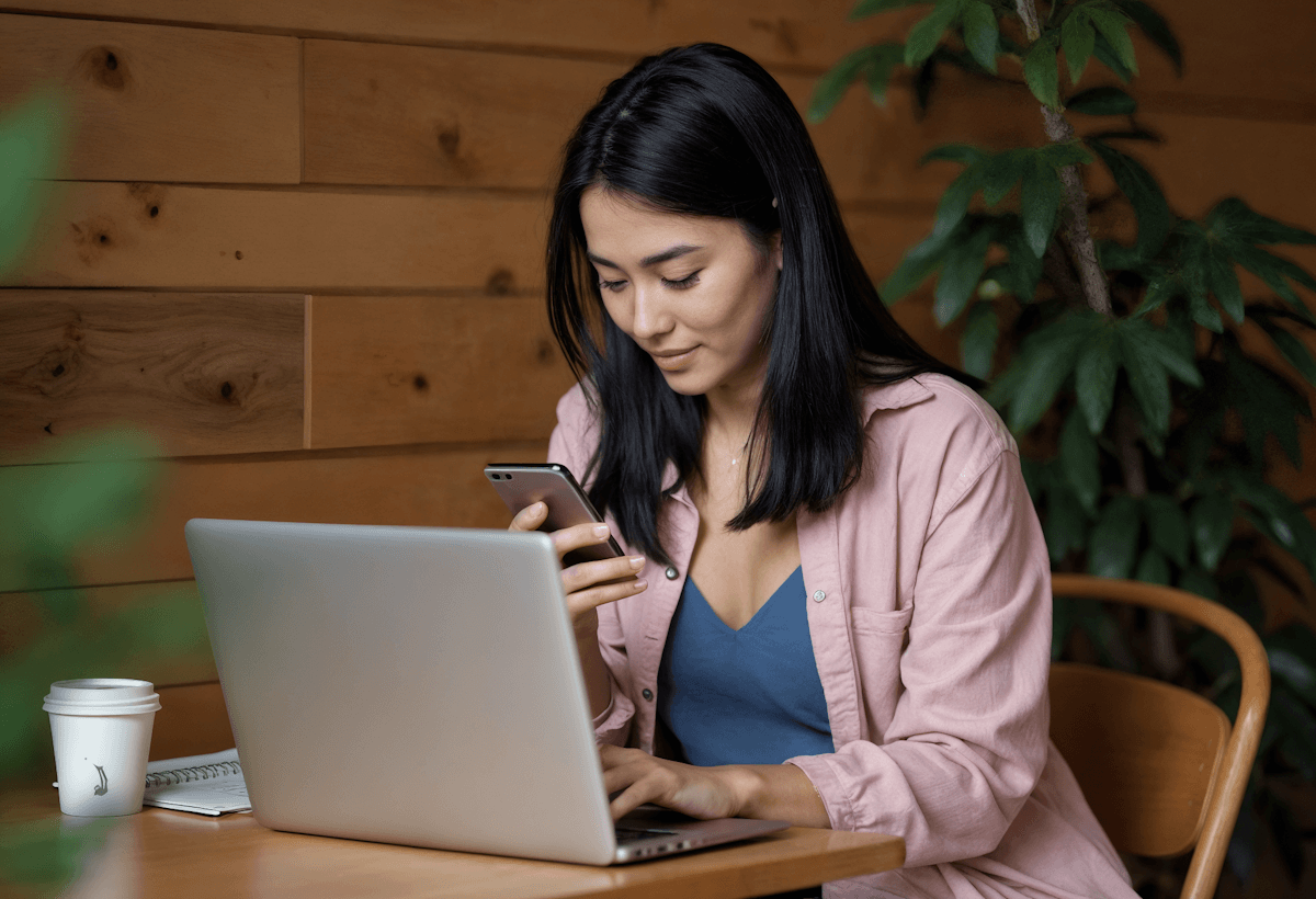 A woman working on her laptop while holding a phone, seated in a cozy setting with a wooden wall and plants in the background