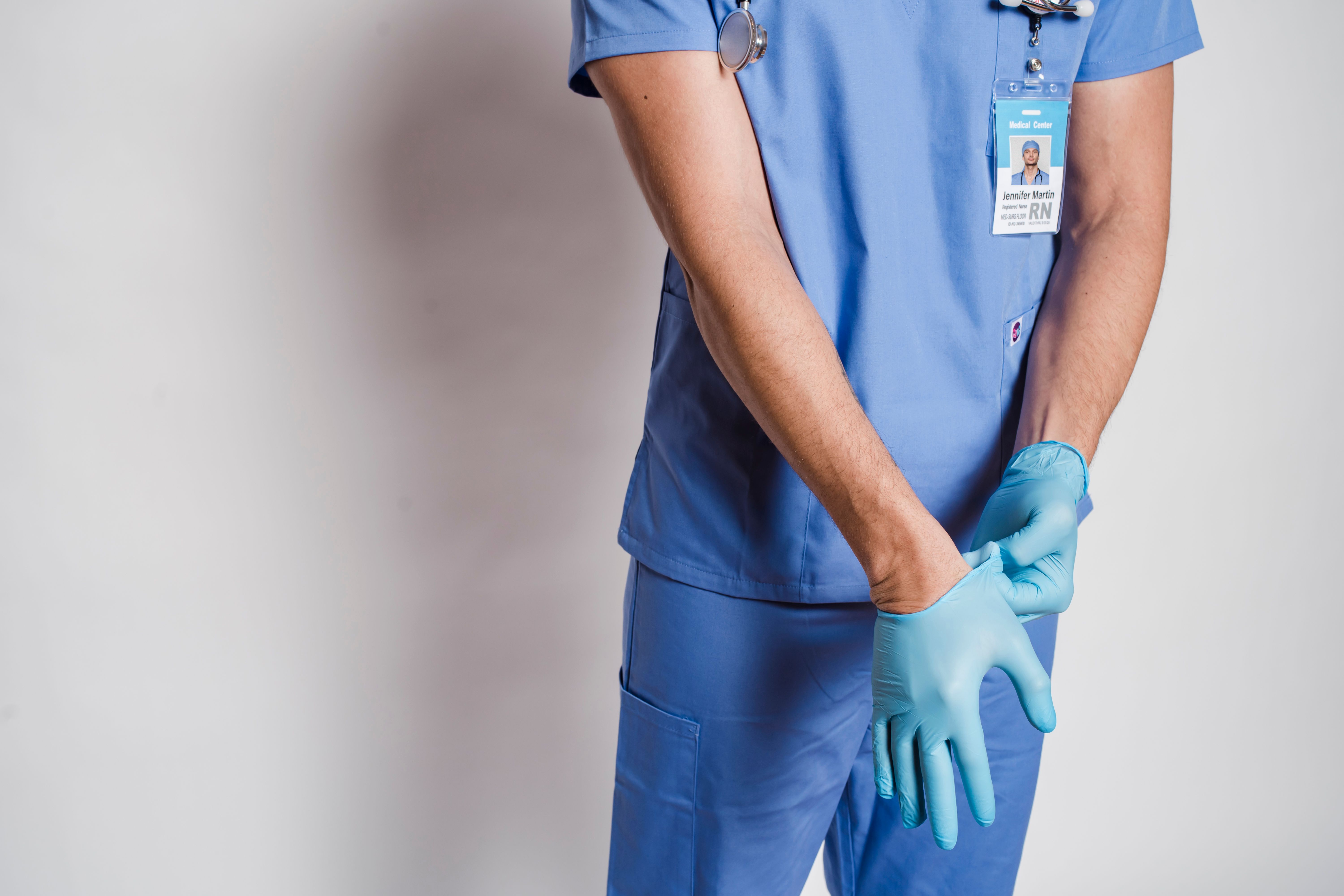 Registered nurse in blue scrubs putting on protective gloves, with a stethoscope and hospital ID badge visible.