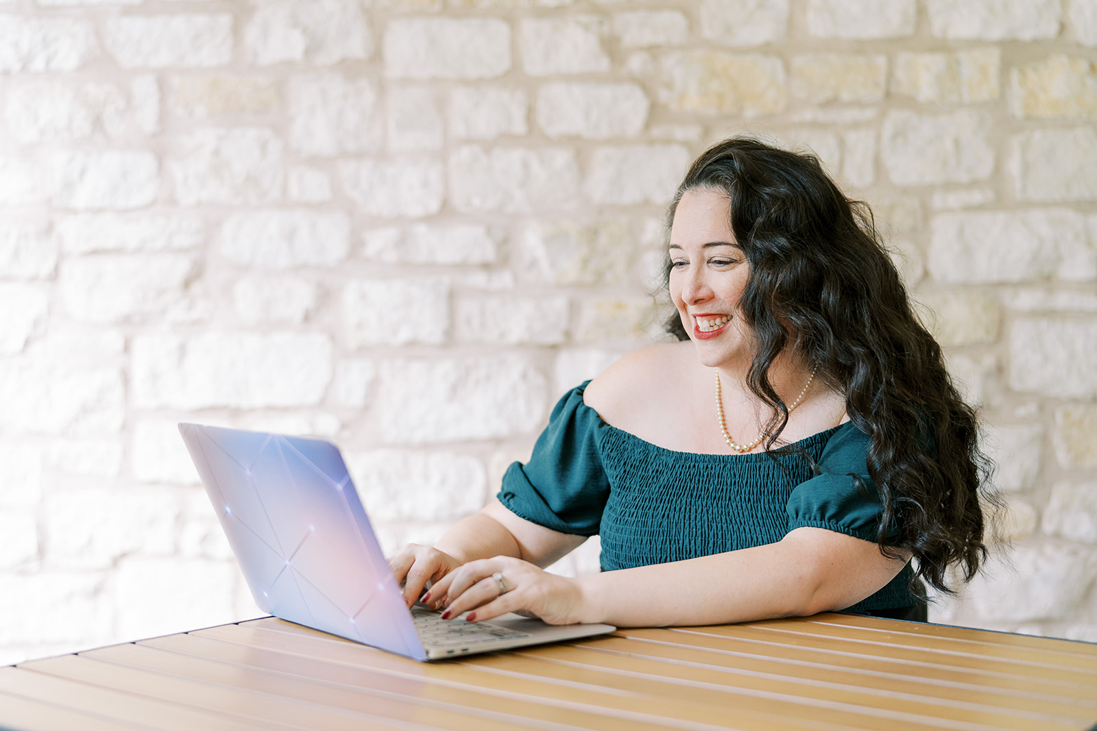 Lizzy Moffett smiling while typing on laptop at wooden desk against brick wall
