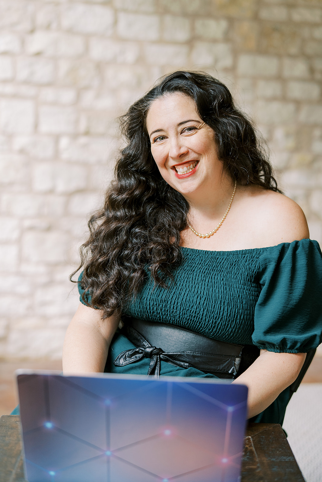Lizzy Moffett smiling at laptop, wearing teal off-shoulder top with pearl necklace