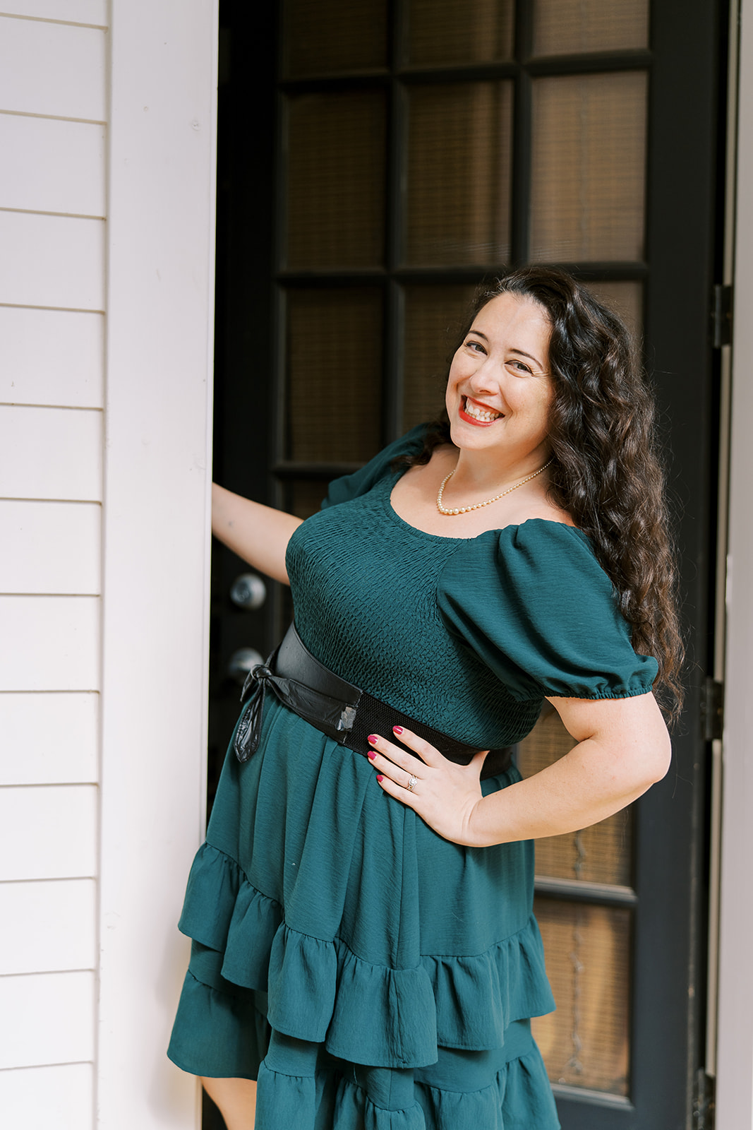 Lizzy Moffett smiling in a teal dress with pearl necklace and black belt.