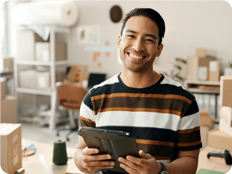 Smiling man holding a tablet in a workspace filled with cardboard boxes and shelves.