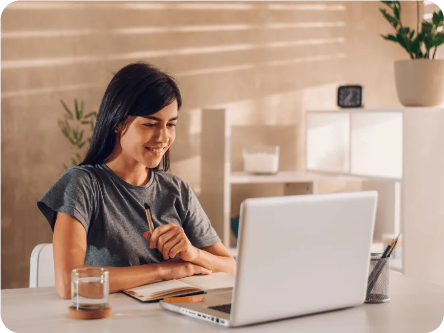 Smiling woman sitting at a desk with a notebook and pen, looking at a laptop screen in a bright room.