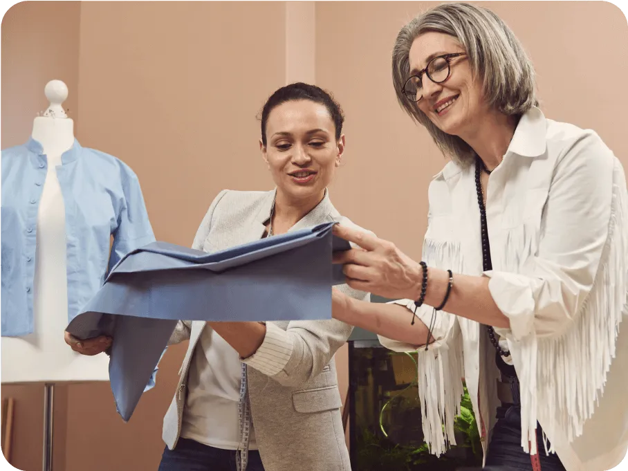 Two women smiling and examining a piece of blue fabric in a sewing or design studio with a dress form in the background.
