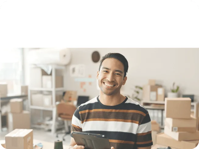 Smiling man holding a tablet in a room filled with cardboard boxes and shelves.