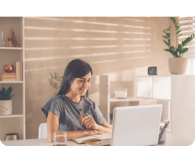 Young woman sitting at desk with laptop, smiling and holding a pen in a warmly lit room.