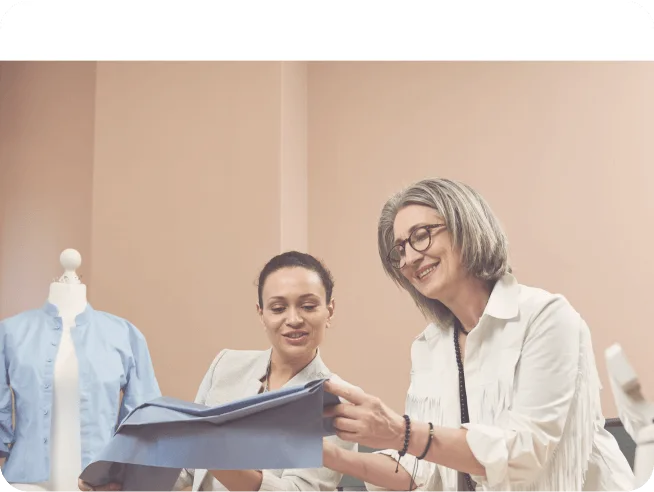 Two women examining blue fabric in a sewing or fashion design studio with a mannequin dressed in a blue garment in the background.