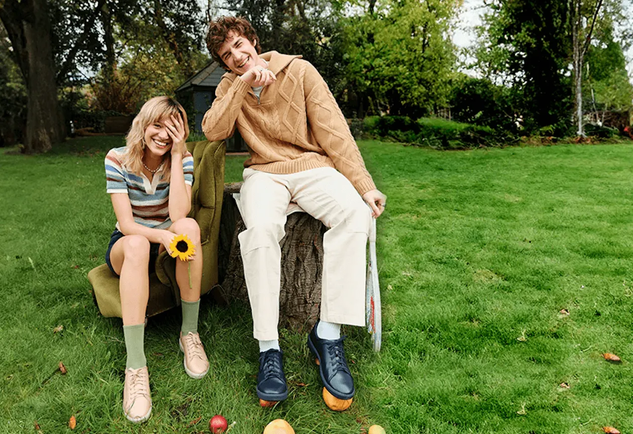 A young man and woman sitting on green grass, the woman holding a sunflower and the man holding a tennis racket, both smiling.