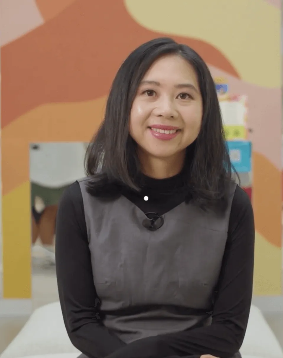 Smiling woman with shoulder-length black hair wearing a grey dress over a black long-sleeve shirt, seated indoors with a colorful abstract wall behind her.