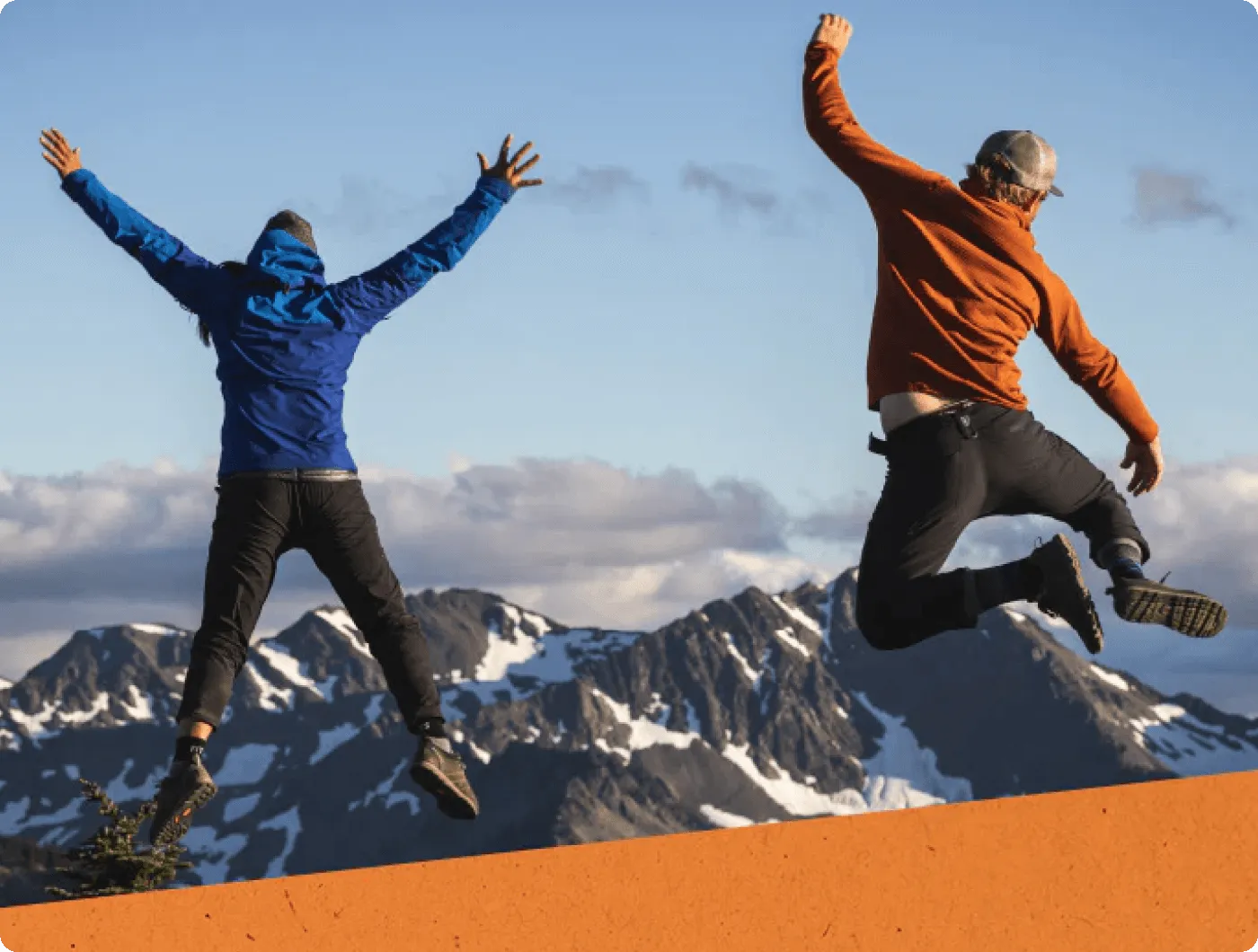 Two people jumping energetically with snowy mountain peaks in the background under a clear sky.