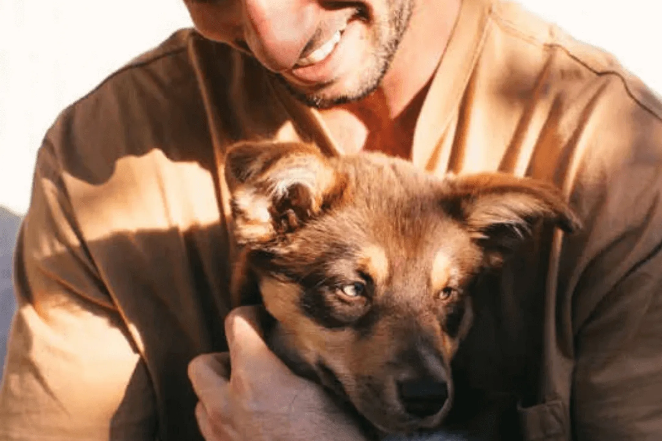 Man smiling and holding a small brown and black puppy close to his chest.