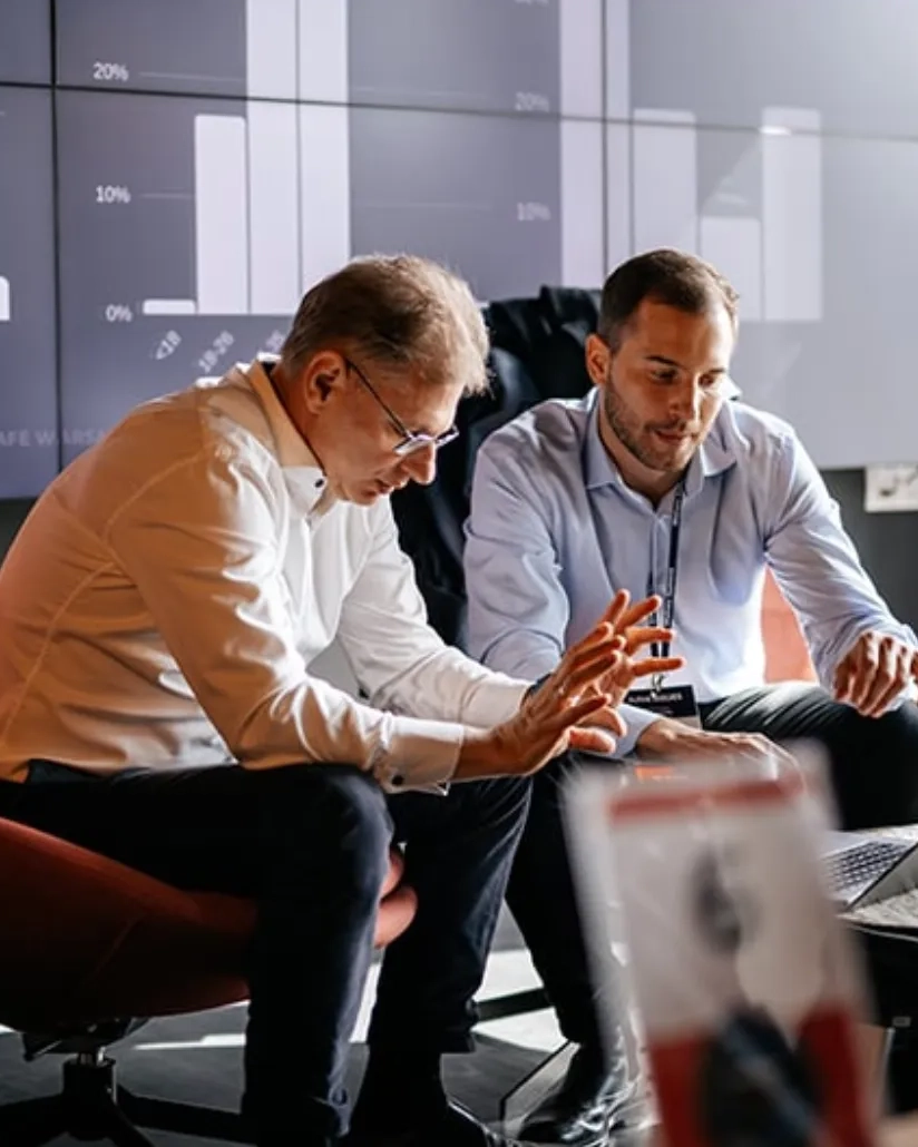 Two men in business attire seated in front of a laptop, discussing something with graphs displayed on large screens in the background.
