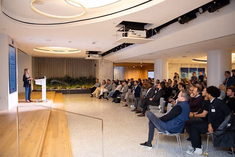 Startup founder presenting a pitch on stage to an audience of investors and venture capital professionals in a modern conference space.