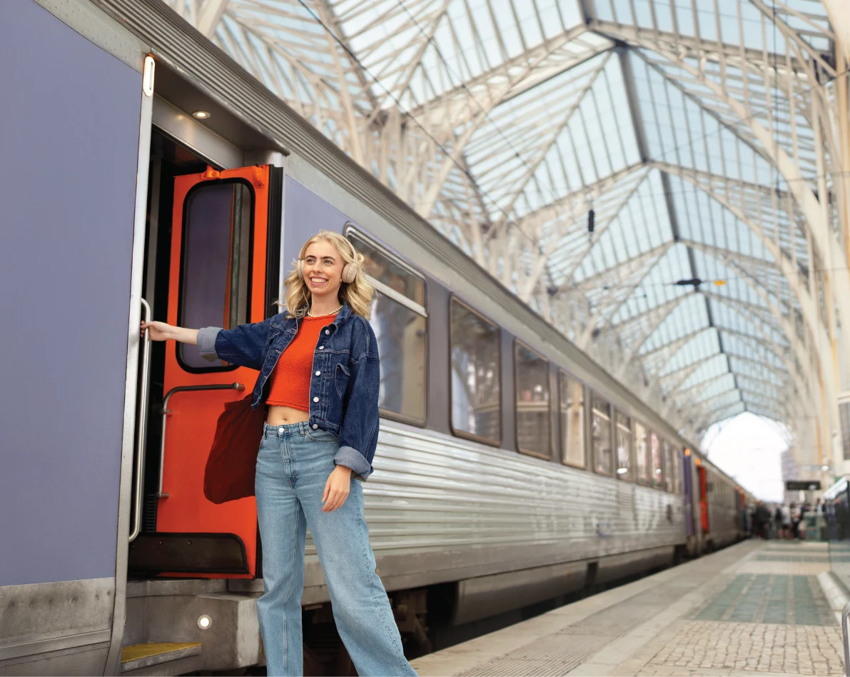 Smiling young woman in denim jacket and orange top standing at open door of train in a large glass-roofed station.