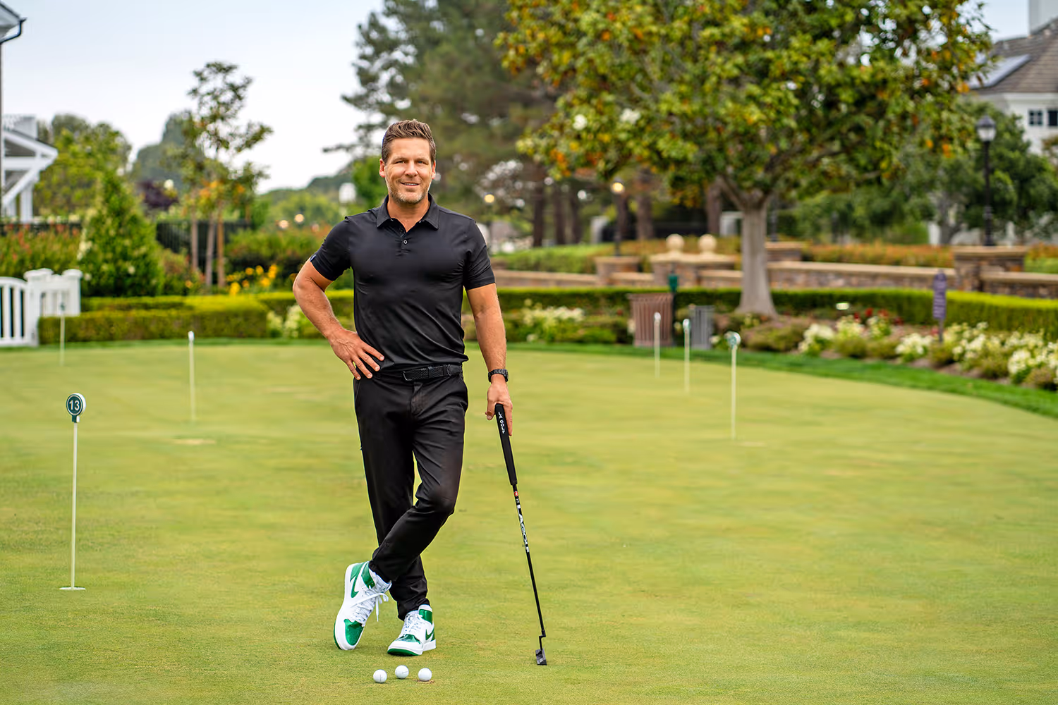 Man in black golf attire standing on a putting green with three golf balls in front and holding a putter.