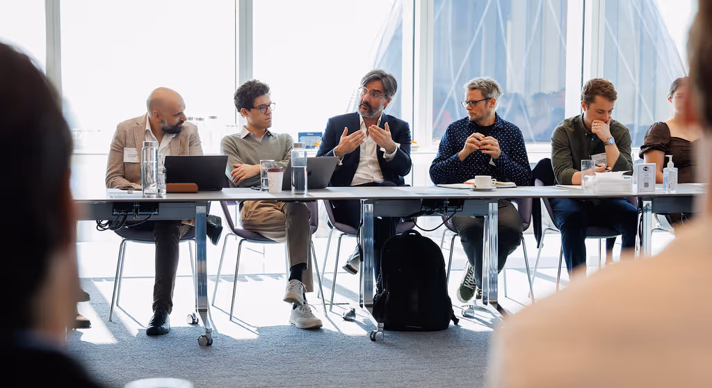 Group of six people seated at a conference table engaged in a discussion during a meeting in a bright office.