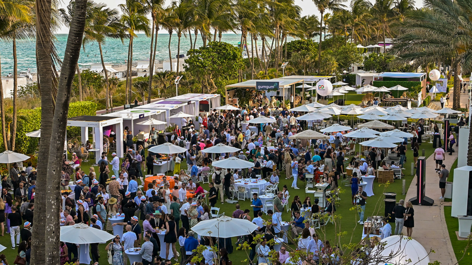Outdoor event with many people seated and standing under white umbrellas, surrounded by palm trees and the ocean in the background.
