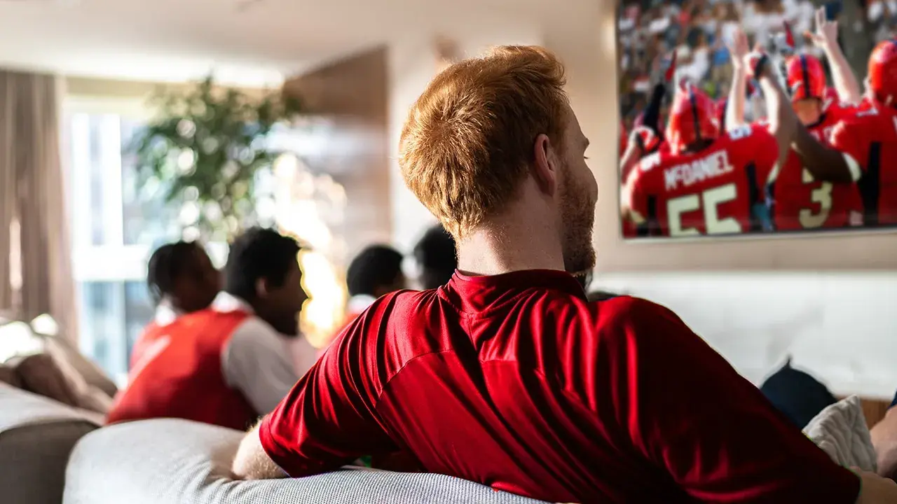Man in red shirt watching American football game on TV with friends in a living room.